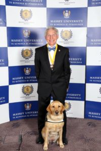 Don Mckenzie and his guid dog, a yellow Labrador named Holly, stand in front of a Massey university sign. Don is wearing a medal around his neck. Holly is sitting bolt upright as if she expects a medal of her own.