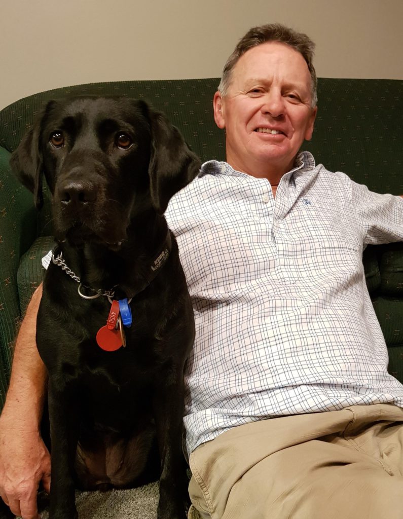 David Wilson sits on a couch with his guide dog, a black Labrador named Cayla